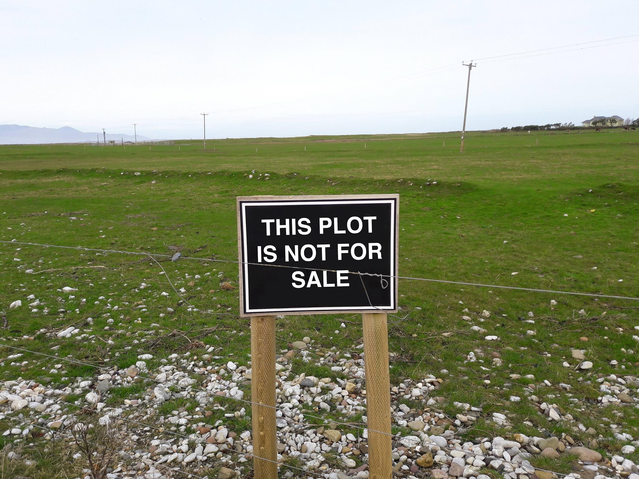 A prominent black sign reading "THIS PLOT IS NOT FOR SALE" in bold white capital letters, securely mounted on wooden posts along a barbed wire fence bordering an open, grassy land parcel.