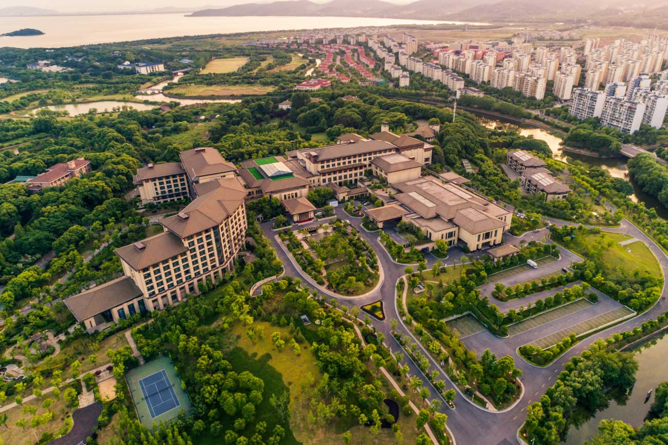 Aerial view of a luxury residential estate and upscale resort complex surrounded by lush green parks, featuring a tennis court, parking amenities, and a distant city skyline during sunset.