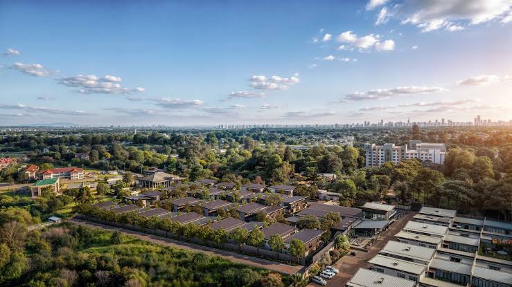 Aerial view of a luxury gated community featuring uniform modern townhouses and villas, set against a lush green suburban landscape with a distant city skyline under a clear blue sky.