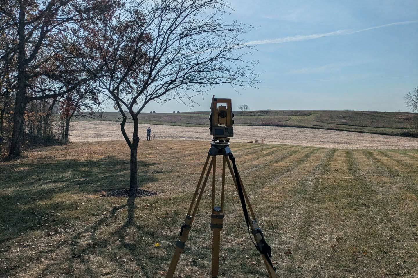 A yellow survey theodolite total station mounted on a wooden tripod in a grassy field, overlooking a large open landscape plot with a surveyor standing in the far distance under a bright sky.