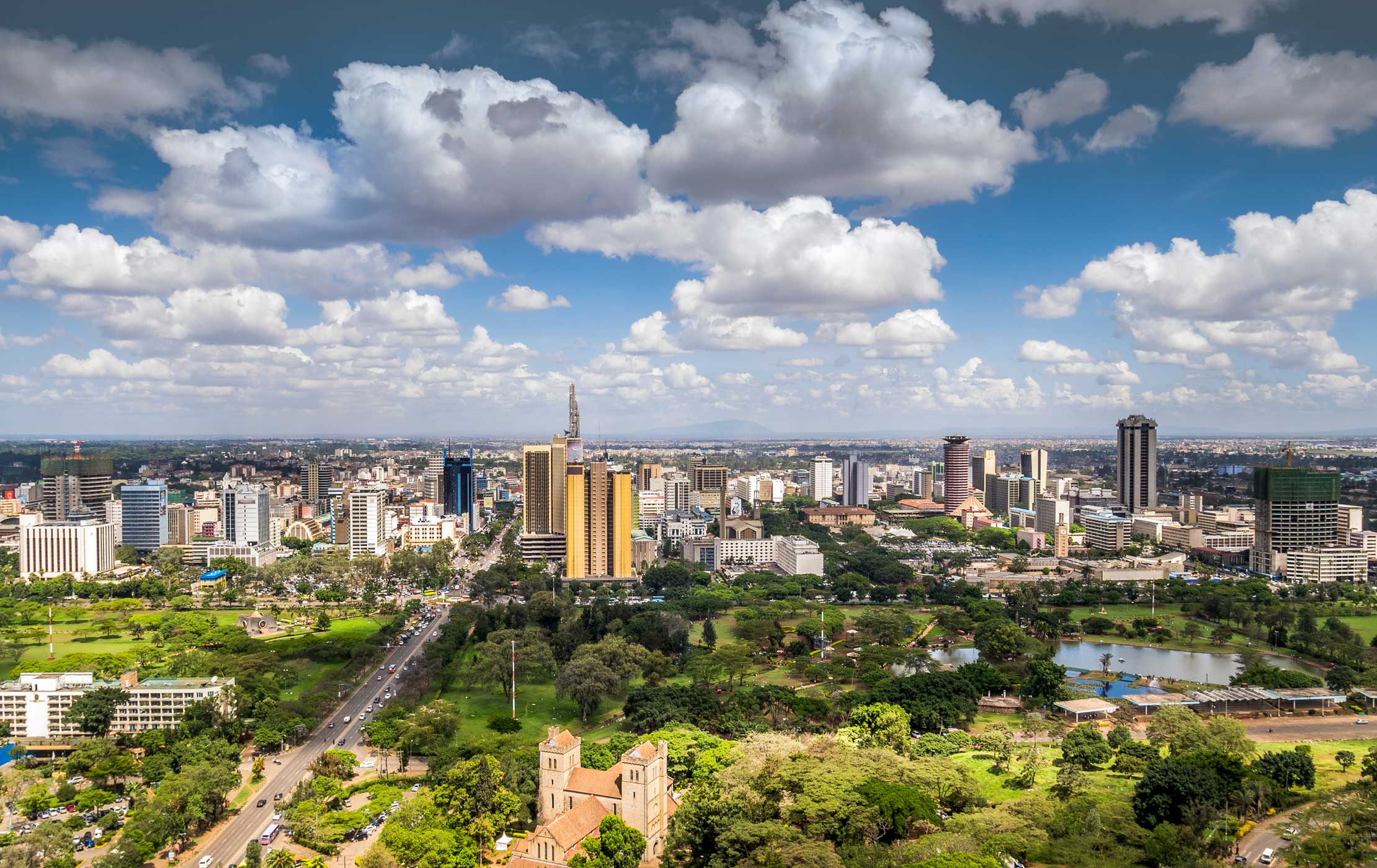 Wide aerial panorama of the Nairobi skyline in Kenya featuring Uhuru Park, the city lake, central business district skyscrapers, and historic stone church architecture under a blue sky.