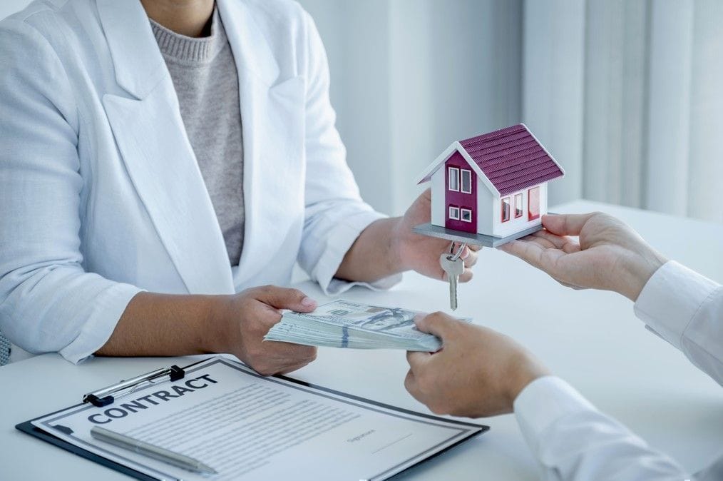 Close-up of a property buyer exchanging cash for a miniature house model and key during a real estate closing transaction, symbolizing land acquisition and property investment.