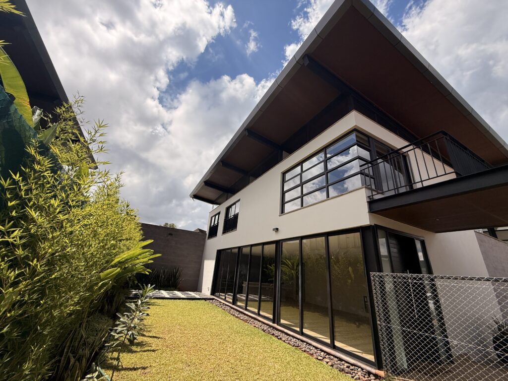 Low-angle view of a modern luxury 4-bedroom townhouse in Runda, Nairobi, featuring a sleek flat-angled roof, expansive floor-to-ceiling glass windows, and a private green lawn garden.