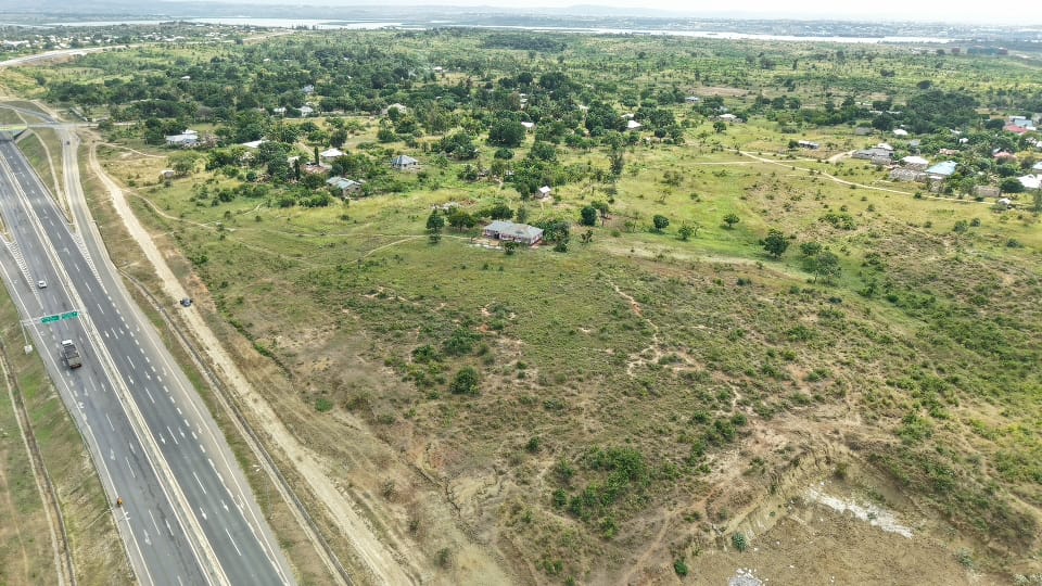 Aerial view of vacant residential and commercial land for sale next to a multi-lane paved highway infrastructure development, showcasing prime plots with rolling green terrain and distant water views.