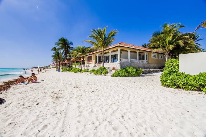 Exterior view of a luxury beachfront villa villa with a tiled roof and glass patio doors, situated directly on a white sand beach with palm trees under a clear blue sky.