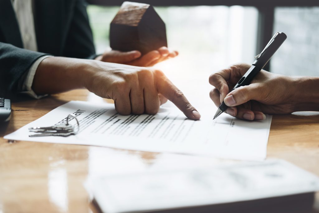 alt="Close-up of a client signing a real estate contract agreement while a property agent points to the signature line and holds a model house, representing a secure land acquisition closing.