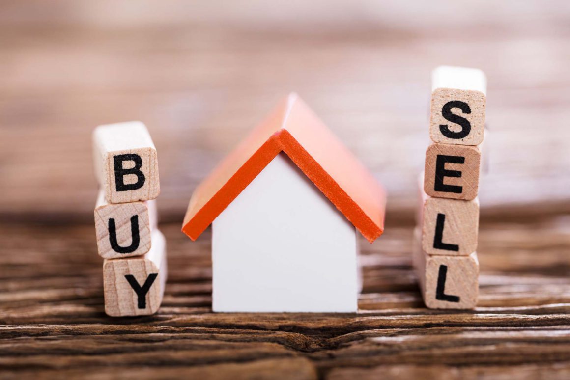 A miniature white toy house with an orange roof positioned between wooden letter blocks spelling Buy and Sell, symbolizing real estate market options, property acquisition, and sales.
