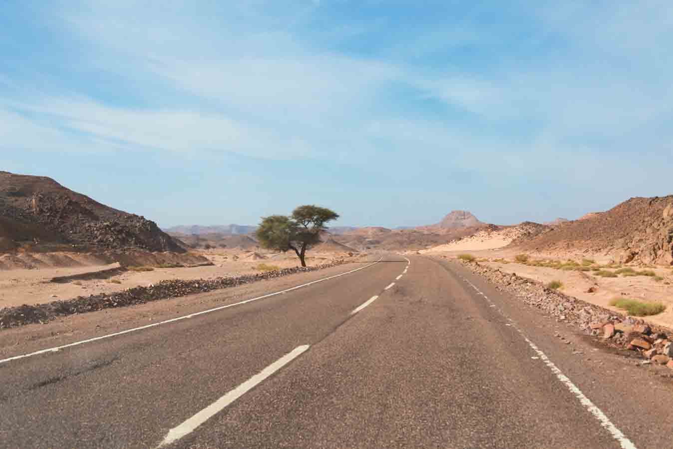 Low-angle view of a paved two-lane asphalt highway passing through a desert mountain landscape, highlighting transit infrastructure that connects remote land plots for acquisition.