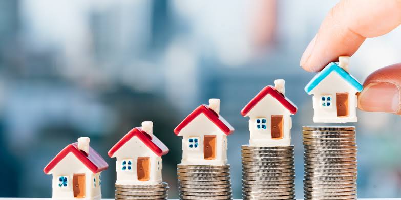 Row of miniature toy houses standing on ascending stacks of silver coins, with a hand placing the final house to symbolize property value growth, real estate ROI, and wealth creation.